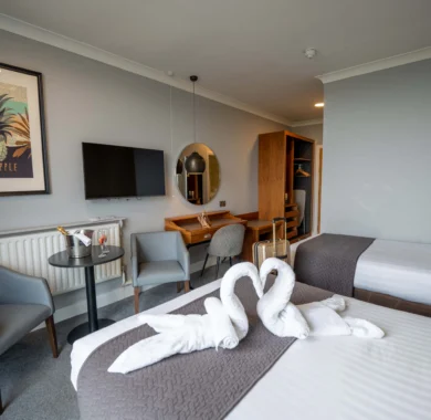 Stylish hotel room with cosy twin beds, swan towels, modern decor, flat-screen TV, workspace, and seating area.