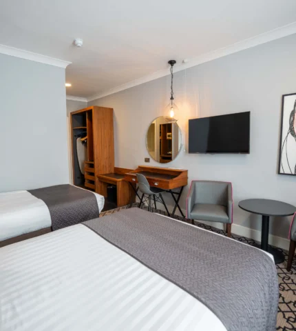 Spacious hotel room with two beds, modern decor, flat-screen TV, cosy chairs, and ample natural light for a relaxing stay.