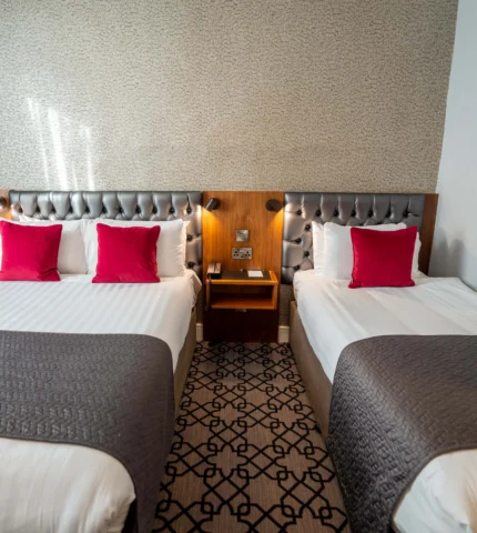 Elegant twin hotel room with plush grey headboards, vibrant red cushions, and modern bedside lamps.